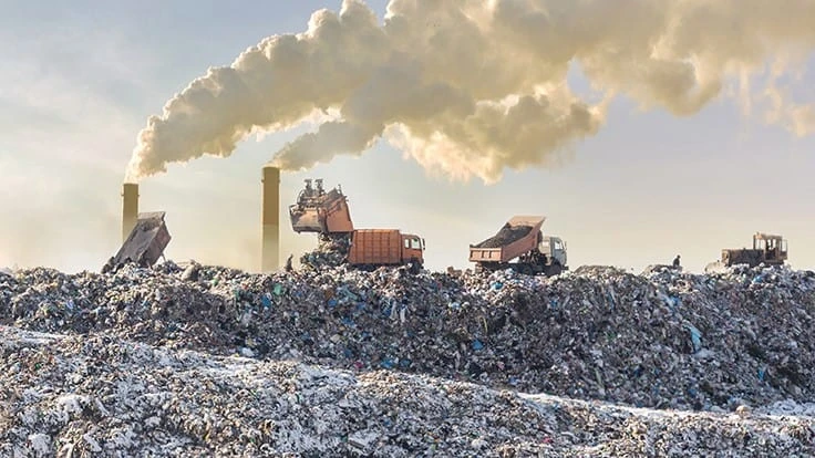 Trucks dumping trash at a landfill with smokestacks venting in the background